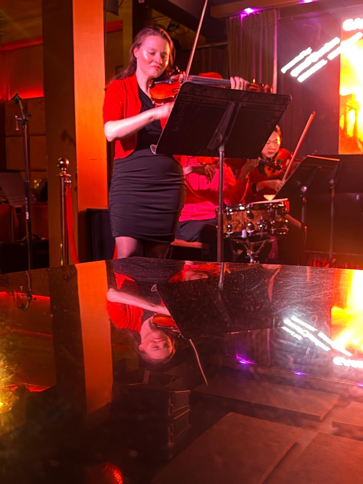 Violinist performing with reflection on piano surface under dramatic red lighting